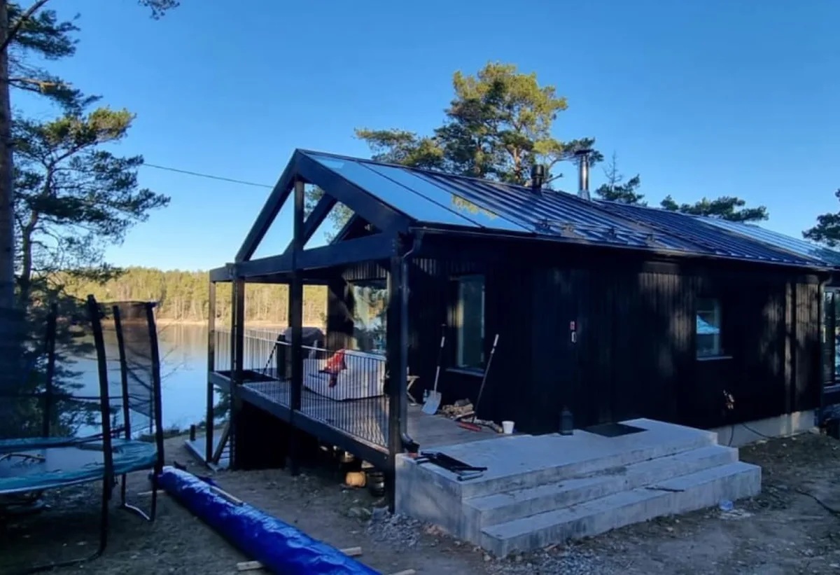 Mörkt trähus med stora glaspartier och altan bland tallar vid vattnet